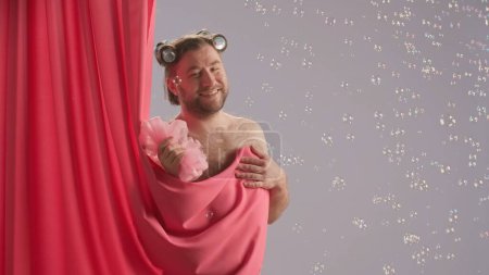 Téléchargez les photos : Un homme avec des bigoudis sur les cheveux regarde derrière un rideau rose et sourit. Homme avec torse nu et coiffé d'un bonnet de douche. Concept de Beauté Humoristique. Les bulles de savon volent autour - en image libre de droit