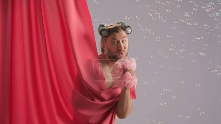 Photo for Funny man with curlers on his head, covered by pink shower curtain singing using loofah as microphone. Comical man showering in studio on blue background surrounded by soap bubbles - Royalty Free Image