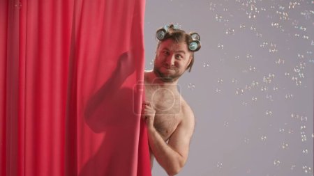 Foto de Un hombre tonteando en la ducha, haciendo caras. Un hombre con rizadores en la cabeza, cubierto con una cortina de ducha rosa en el estudio sobre un fondo azul rodeado de burbujas de jabón de cerca - Imagen libre de derechos