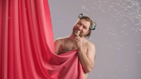 Téléchargez les photos : Un homme drôle avec des bigoudis sur la tête, recouvert d'un rideau de douche rose, chante en utilisant une brosse à dents comme microphone. Homme comique se douchant en studio sur fond bleu entouré de bulles de savon - en image libre de droit