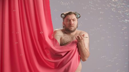 Téléchargez les photos : L'homme, couvert d'un rideau de douche rose, hausse les épaules confusément et écarte les mains. Un homme avec un bandeau sur la tête et une brosse à dents à la main entourée de bulles de savon dans la - en image libre de droit