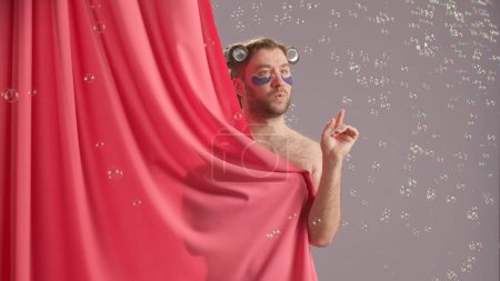 Foto de Seminude hombre cubierto por una cortina de ducha rosa estallando burbujas de jabón girando a su alrededor. Hombre divertido con rizadores en la cabeza y parches de hidrogel bajo los ojos en el estudio sobre fondo azul - Imagen libre de derechos