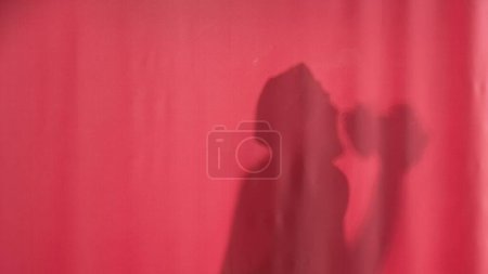 Téléchargez les photos : Silhouette d'un homme se lavant derrière un rideau rose. Homme au bonnet imperméable à l'eau lavant, dansant et chantant, utilisant loofah comme microphone. Lavage corporel - en image libre de droit