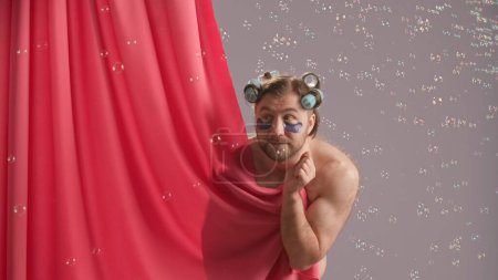 Foto de Seminude hombre cubierto con cortina de ducha rosa en el estudio sobre fondo azul rodeado de burbujas de jabón. Un hombre con rulos en la cabeza y manchas de hidrogel bajo sus ojos inclina su dedo hacia su boca - Imagen libre de derechos