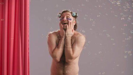 Téléchargez les photos : Un homme en bigoudis et avec des taches d'hydrogel sous les yeux dit wow dans l'excitation et fait un geste de victoire avec ses mains. Homme heureux en studio près du rideau de douche rose entouré de bulles de savon - en image libre de droit