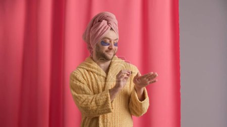 Photo for A man is grooming his nails after a shower, applying nail polish, nail oil with a brush. Man in yellow robe, with towel on his head and with hydrogel patches under his eyes in studio against pink - Royalty Free Image