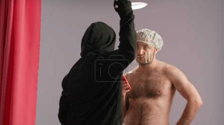 Foto de Un maníaco con una máscara blanca con un cuchillo ataca a un hombre con un torso desnudo bañándose en la ducha. Un hombre con una gorra impermeable mira fijamente al maníaco - Imagen libre de derechos
