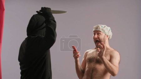 Foto de Un maníaco con una máscara blanca con un cuchillo ataca a un hombre con un torso desnudo bañándose en la ducha. Un hombre con gorra impermeable mira al maníaco y grita de horror - Imagen libre de derechos