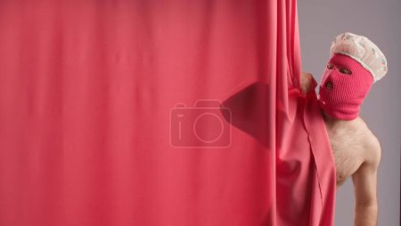 Foto de Un hombre con un torso desnudo con un pasamontañas rosa y una gorra de ducha asoman por detrás de una cortina rosa. Un hombre en una imagen divertida tomando una ducha - Imagen libre de derechos