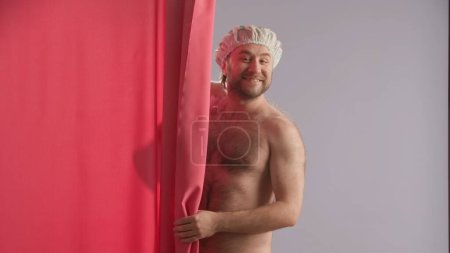 Foto de Un hombre con un torso desnudo se asoma por detrás de una cortina de ducha rosa y sonríe. Un hombre que lleva una gorra de ducha está realizando procedimientos de higiene. Concepto de belleza humorístico - Imagen libre de derechos