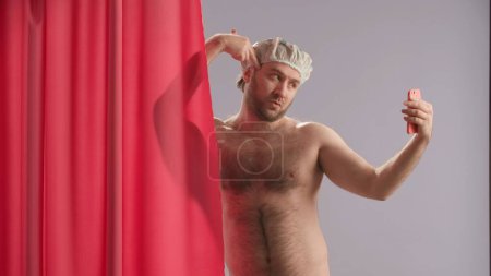 Foto de Un hombre se toma selfies usando un teléfono inteligente mientras se ducha. Hombre desnudo con gorra impermeable, cerca de la cortina de ducha rosa posa en el estudio sobre fondo azul de cerca - Imagen libre de derechos