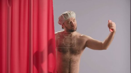 Photo for A man takes selfies using a smartphone while showering. Nude man wearing waterproof cap, near pink shower curtain poses in studio on blue background close up - Royalty Free Image