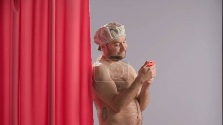 Photo for Man in waterproof shower cap in studio on blue background using smartphone. Man standing near pink shower curtain with a dreamy look typing a message, browsing social media, smiling sending an air - Royalty Free Image