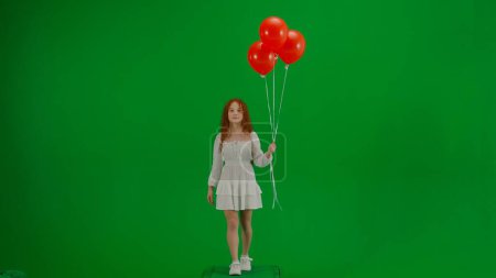 Téléchargez les photos : Concept créatif pour enfants. Portrait de petite fille en studio. Petite fille en robe blanche avec ballons rouges sur écran vert chroma clé isolé marche en regardant la caméra, visage positif. - en image libre de droit