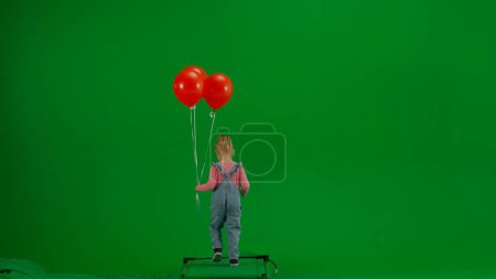 Téléchargez les photos : Concept créatif pour enfants. Portrait de petite fille en studio. Petite fille en jeans dans l'ensemble et queue de cheval avec des ballons d'hélium rouge marchant sur écran vert chromatique isolé, vue de dos. - en image libre de droit