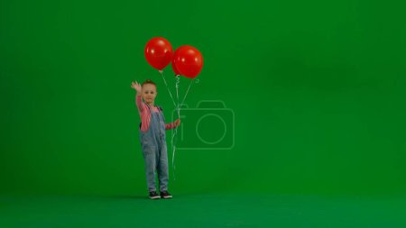 Téléchargez les photos : Une petite fille en salopette denim avec un ballon rouge regarde la caméra et salue les vagues. L'enfant est sur un fond vert chrome clé. Concept de vacances, anniversaire, joie - en image libre de droit