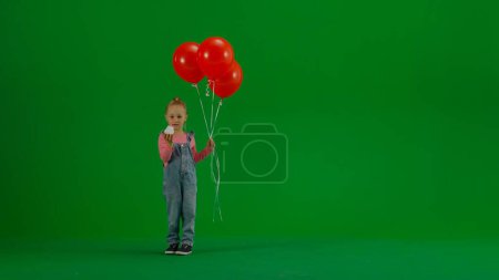 Foto de Niña en overoles de mezclilla con globos rojos mira a la cámara y muestra una magdalena de cumpleaños con vela. Bebé en clave croma fondo verde. Concepto de vacaciones, cumpleaños, alegría - Imagen libre de derechos
