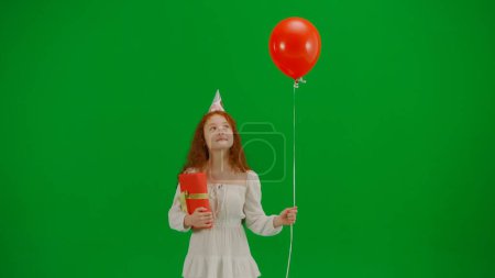 Foto de Concepto creativo para niños. Retrato de niña pequeña en el estudio. Niña en vestido blanco con globo rojo croma clave pantalla verde aislado fondo en sombrero de fiesta con caja de regalo caminando mirando a su alrededor. - Imagen libre de derechos