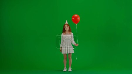 Foto de Concepto creativo para niños. Retrato de niña pequeña en el estudio. Niña en vestido blanco con globo de helio rojo en croma pantalla verde clave fondo aislado en partido sombrero cono mirando a la cámara. - Imagen libre de derechos