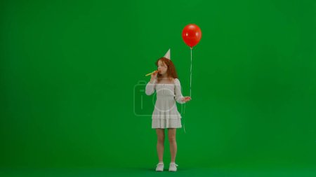 Photo for Children creative concept. Portrait of small girl in studio. Little girl in white dress with red helium balloon on chroma key green screen isolated background in party cone hat blowing horn blower. - Royalty Free Image
