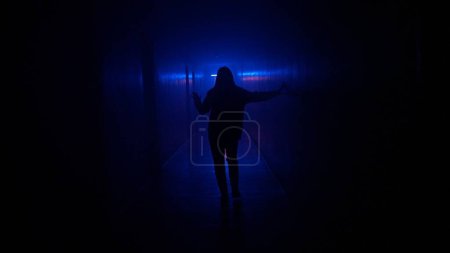 Photo for Abandoned places and empty buildings creative advertisement concept. Portrait of female in hallway blue neon light. Close back shot woman walking cautiously in the dark corridor, holding up to wall. - Royalty Free Image