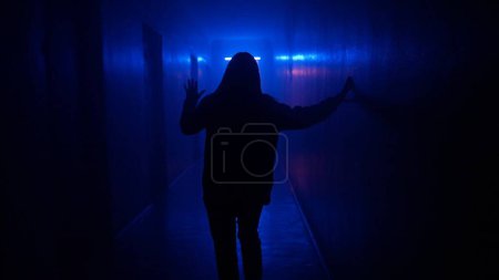 Téléchargez les photos : Lieux abandonnés et bâtiments vides concept publicitaire créatif. Portrait de femme dans le couloir bleu néon lumière. Gros plan arrière femme marchant prudemment dans le couloir sombre, tenant jusqu'au mur. - en image libre de droit
