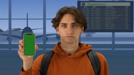 Photo for In the close-up shot of the airport with the waiting room. A man in the foreground looks into the camera, smiles and holds the phone green screen to the camera. Advertising space. - Royalty Free Image