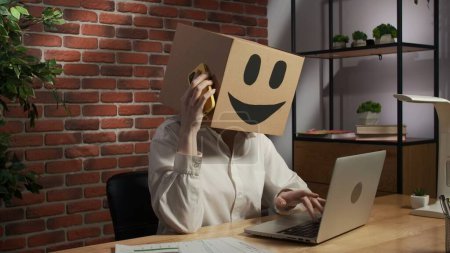 Photo for Business life and office daily routine creative advertisement concept. Portrait of female in cardboard box with emoji on head. Worker sitting at the desk working on laptop, talking on smartphone. - Royalty Free Image