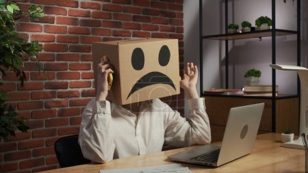 Photo for Business life and office daily routine creative advertising concept. Portrait of a woman in a cardboard box with a negative emoji on her head. Employee sitting at desk, working on laptop, talking on - Royalty Free Image