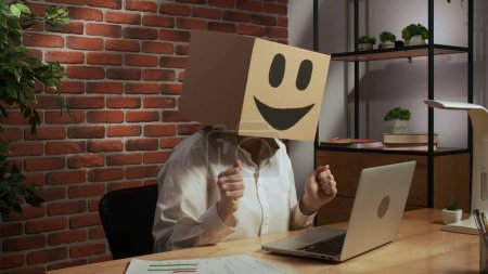 Photo for Portrait of a woman in a cardboard box with a smiling smiley face on her head. Employee at desk working on laptop - Royalty Free Image