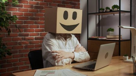 Photo for Portrait of a woman in a cardboard box with a smiling smiley face on her head. Employee at desk working on laptop, sitting cross-armed - Royalty Free Image