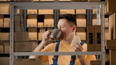 Téléchargez les photos : Entrepôt d'affaires et logistique concept publicitaire créatif. Portrait d'un homme travaillant dans un entrepôt. Homme magasinier debout près de rack manger sandwich boire du café à partir d'une tasse ayant pause déjeuner. - en image libre de droit
