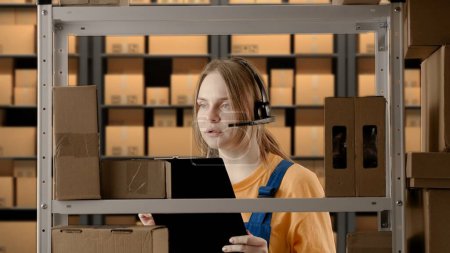 Photo for Business warehouse and logistics creative advertisement concept. Portrait of female model working in storage. Girl storekeeper in uniform standing near rack with boxes, talking in headset checks goods - Royalty Free Image
