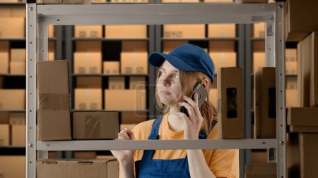 Photo for Business warehouse and logistics creative advertisement concept. Portrait of female model working in storage. Girl storekeeper in uniform near rack with boxes, talking on smartphone, neutral face. - Royalty Free Image
