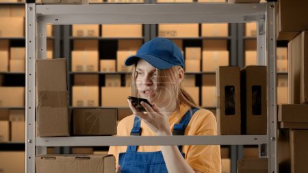 Photo for Business warehouse and logistics creative advertisement concept. Portrait of female model working in storage. Girl storekeeper in uniform near rack with boxes, talking on smartphone, neutral face. - Royalty Free Image