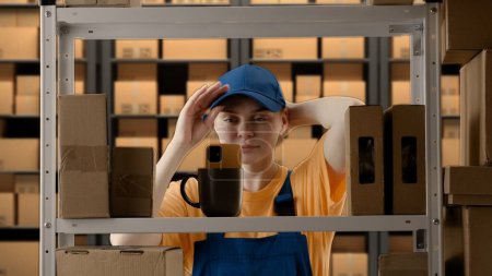 Photo for Business warehouse and logistics creative advertisement concept. Portrait of female model working in storage. Girl storekeeper in uniform near rack with boxes, fixing look taking selfie on smartphone. - Royalty Free Image