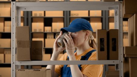 Photo for Business warehouse and logistics creative advertisement concept. Portrait of female model working in storage. Girl storekeeper in uniform standing near rack with boxes, having break drinking coffee. - Royalty Free Image