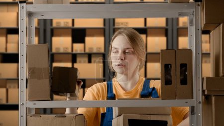 Photo for Business warehouse and logistics creative advertising concept. Portrait of a female model working in a warehouse. Uniformed warehouse girl lays on a shelf cardboard box - Royalty Free Image