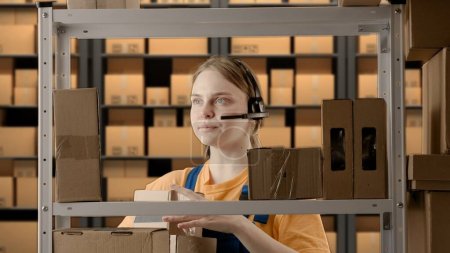 Photo for Business warehouse and logistics creative advertisement concept. Portrait of female model working in storage. Girl storekeeper in uniform near rack with boxes talking in headset organizing shelf. - Royalty Free Image