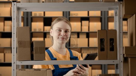 Photo for Business warehouse and logistics creative advertisement concept. Portrait of female model working in storage. Girl storekeeper in uniform standing near rack with boxes, checking goods on tablet. - Royalty Free Image