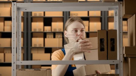 Photo for Business warehouse and logistics creative advertising concept. Portrait of a female model working in a warehouse. Uniformed warehouse girl lays on a shelf cardboard box - Royalty Free Image