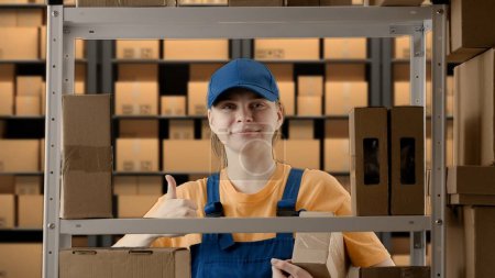Photo for Business warehouse and logistics creative advertisement concept. Portrait of female model working in storage. Girl storekeeper in uniform standing near rack with boxes, takes package shows thumbs up. - Royalty Free Image