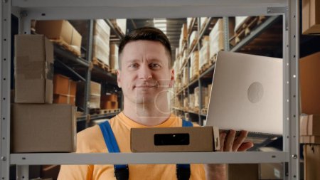 Photo for Business warehouse and logistics creative advertisement concept. Portrait of male model working in storage. Man storekeeper standing near rack with boxes, checking goods, typing information on laptop. - Royalty Free Image