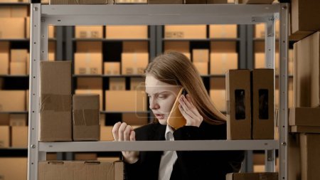 Photo for Business warehouse and logistics creative advertisement concept. Portrait of female working in storage. Girl storekeeper manager in formal outfit near rack with boxes, talking on smartphone. - Royalty Free Image