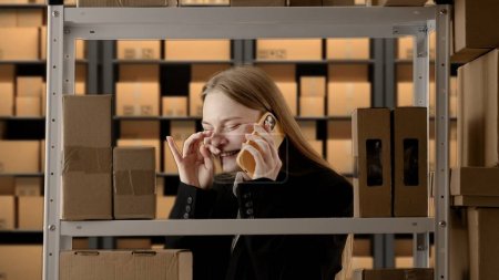 Photo for Business warehouse and logistics creative advertisement concept. Portrait of female working in storage. Girl storekeeper manager in formal outfit near rack with boxes, talking laughing on smartphone. - Royalty Free Image