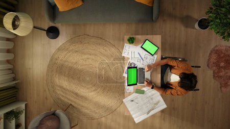 Photo for Top view of a disabled young man on a wheelchair. Architect is working on a laptop and a tablet with an advertising area, workspace mock up on them, studying the blueprints. Remote, modern lifestyle. - Royalty Free Image