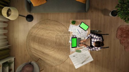 Photo for Top view of a disabled young woman on a wheelchair. Architect working on a laptop and a tablet with an advertising area, workspace mock up on them, studying the blueprints. Remote, modern lifestyle. - Royalty Free Image