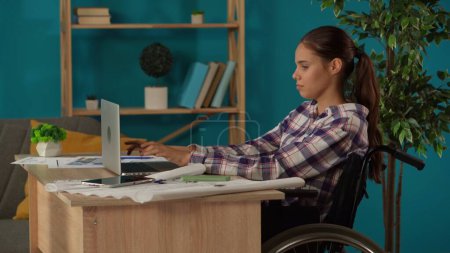 Foto de Un disparo medio capturando a una joven discapacitada en una silla de ruedas que trabaja en su oficina en casa. Joven arquitecto mirando a través de los planos y deslizando en una pantalla de tableta. Trabajo remoto, estilo de vida moderno. - Imagen libre de derechos