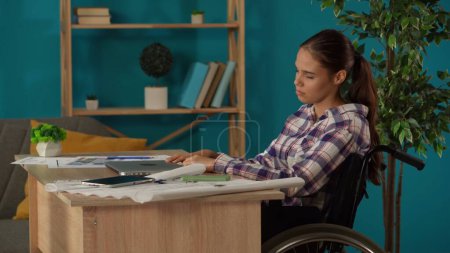 Photo for Medium shot capturing a disabled young woman on a wheelchair working in her home office. Young architect looking through the blueprints and swiping on a tablet screen. Remote job, modern lifestyle. - Royalty Free Image