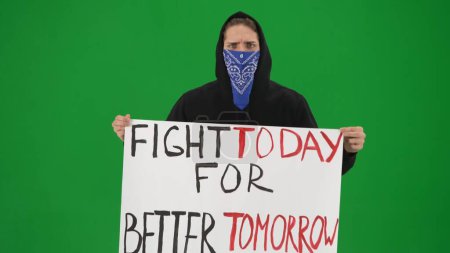 Photo for A male activist holds a placard that reads Fight today for a better tomorrow. A man in a hoodie with his face covered by a scarf on a green screen, close up. Protest. Motivation. Rally. Future - Royalty Free Image